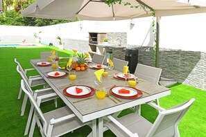 An outdoor dining area with a table set for six, surrounded by white chairs, under a large umbrella.