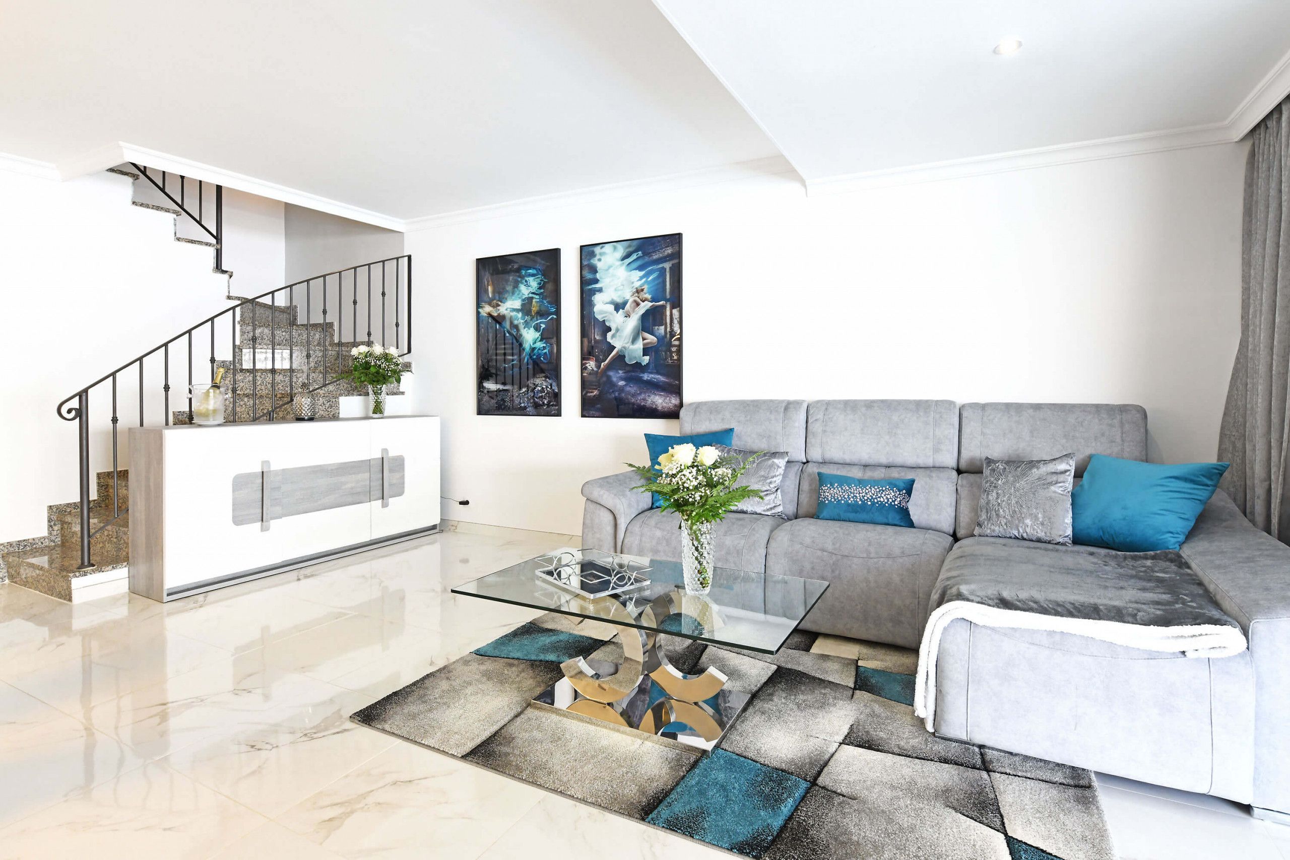 Un salon moderne avec une table basse en verre, un canapé gris agrémenté de coussins bleus, un escalier et deux œuvres d’art accrochées au mur.
