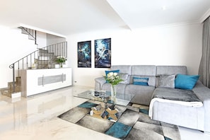 A modern living room with a glass coffee table, grey sofa with blue cushions, a staircase, and two wall-mounted artworks.