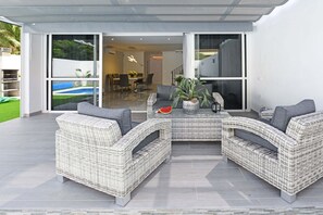 A modern outdoor living area with a wicker sectional sofa, a glass door leading to a dining area, and a small table with a plant.