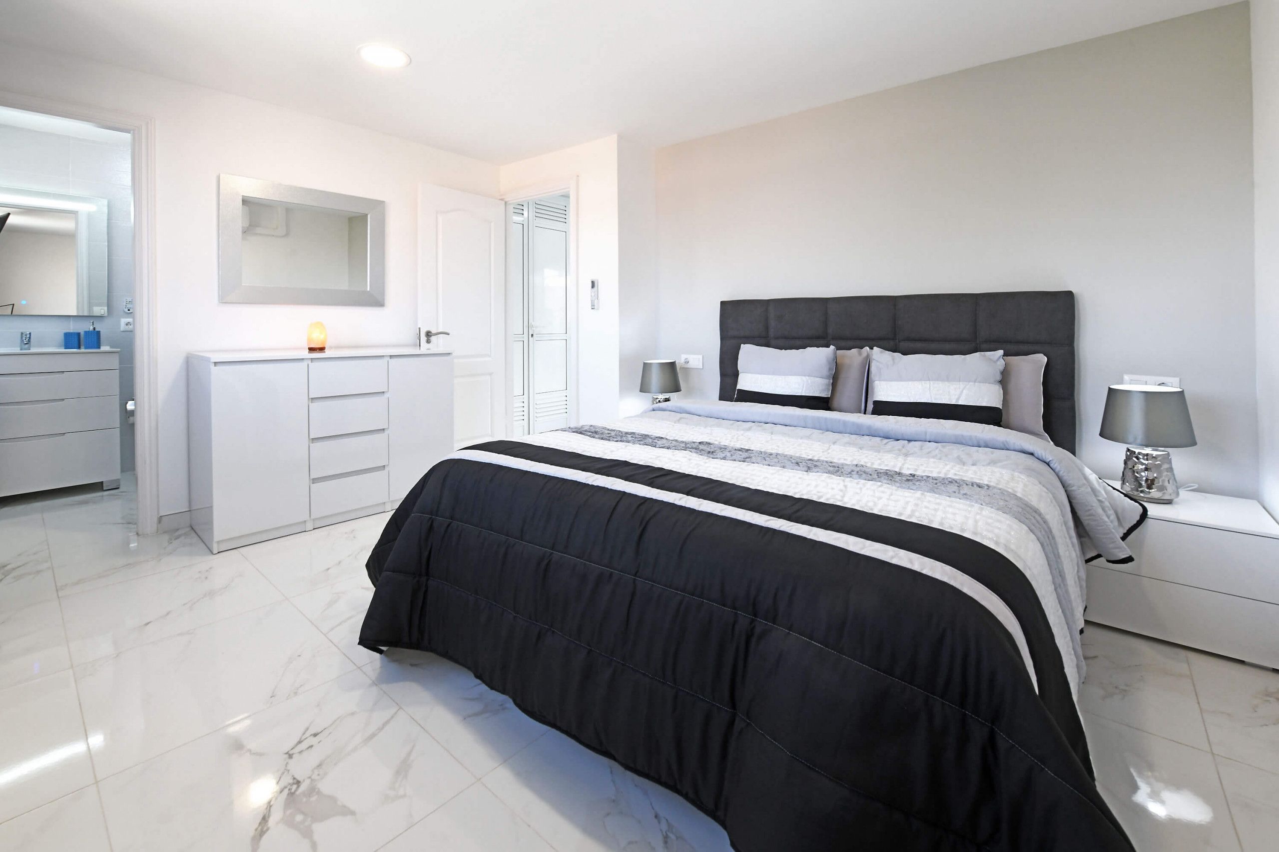 Un dormitorio moderno con una cama grande, una mesita de noche con lámpara, un armario y un espejo.
