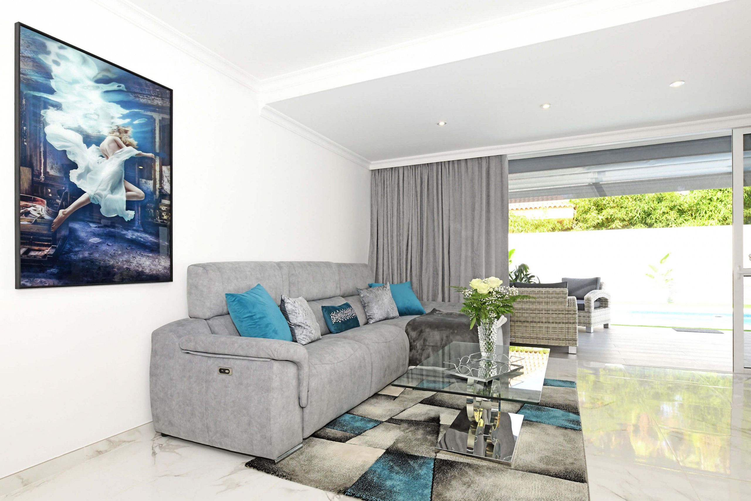 A modern living room with a grey sofa, blue cushions, a glass coffee table, and a large painting.