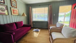 Quarto com piso de madeira, um sofá vermelho, um sofá branco e uma mesa pequena. Há quadros nas paredes e uma janela com cortinas.