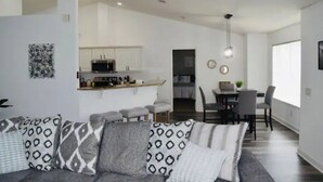 Una sala moderna con un sofá gris, cojines estampados, un comedor con mesa y sillas, y una cocina al fondo.