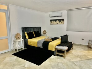 A bedroom with a bed, bedside tables, a bedside lamp, a small stool, a black rug, and a wall-mounted air conditioner.