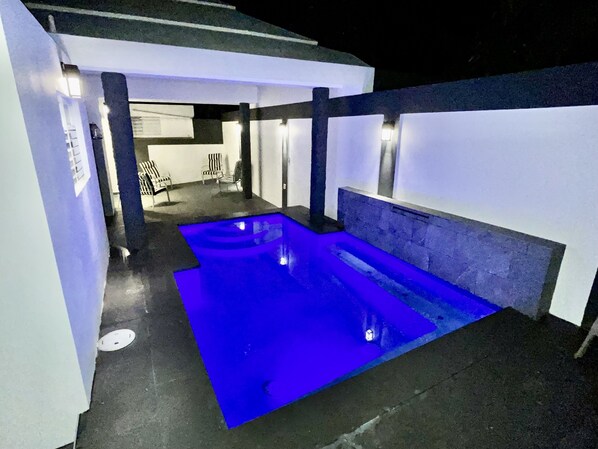 A modern outdoor pool area with a blue-lit pool, surrounded by white walls and black pillars.