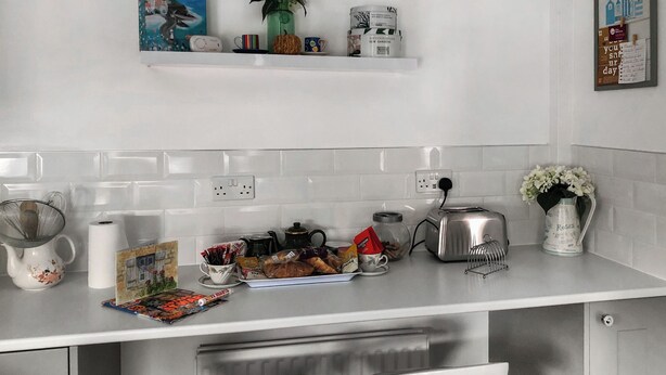 Een moderne keuken met witte aanrechtbladen, een koffiezetapparaat, een broodrooster en een waterkoker. Er zijn twee hangende hanglampen, een plank met decoratieve objecten, en een lijst met een afbeelding aan de muur.