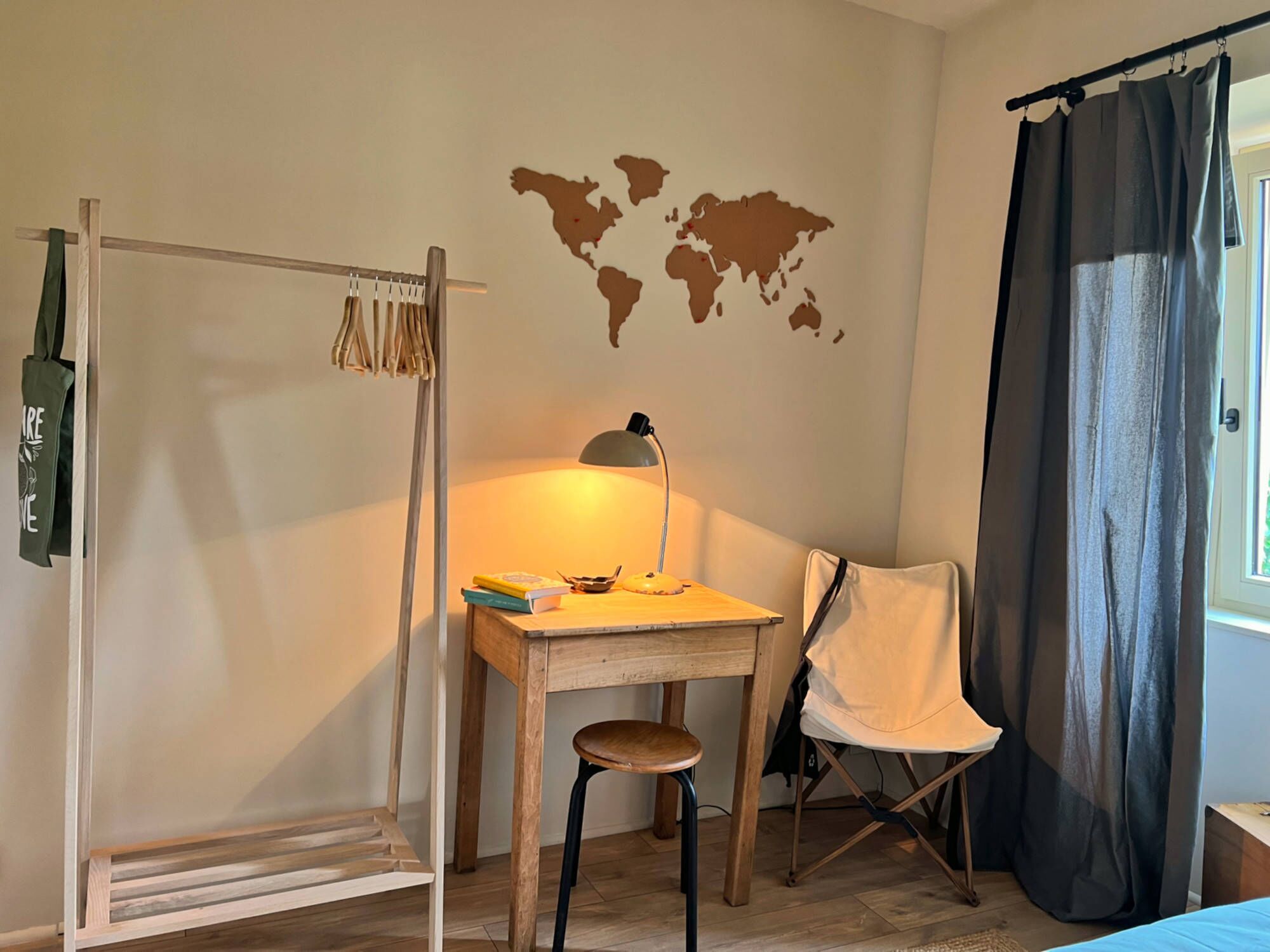 Une pièce avec un bureau en bois, une chaise, un porte-manteau suspendu, une carte du monde accrochée au mur et une fenêtre avec des rideaux.