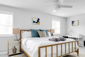 Un dormitorio moderno con cama de madera, ropa de cama blanca, almohadas verde azulado y color beige, un escritorio y obras de arte en las paredes.