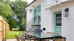 Eine Terrasse im Freien mit Tischplatte aus Glas und Korbstühlen, ein Holzzaun und ein weißes Gebäude mit grüner Tür und Fenster.