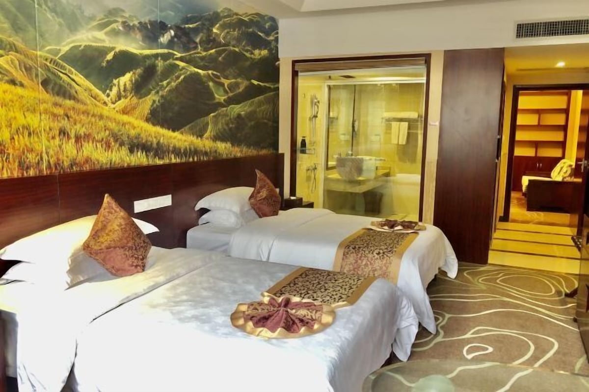 Habitación de hotel con dos camas, un mural grande de un paisaje montañoso, un baño con paredes de vidrio y una escalera de madera.