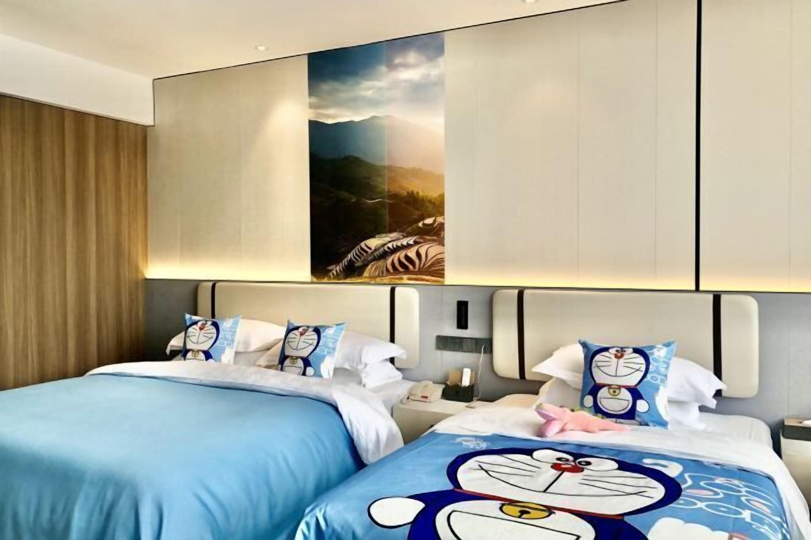 A hotel room with two beds, each with a blue bedspread featuring cartoon characters. There is a large painting above the beds depicting a mountain landscape.
