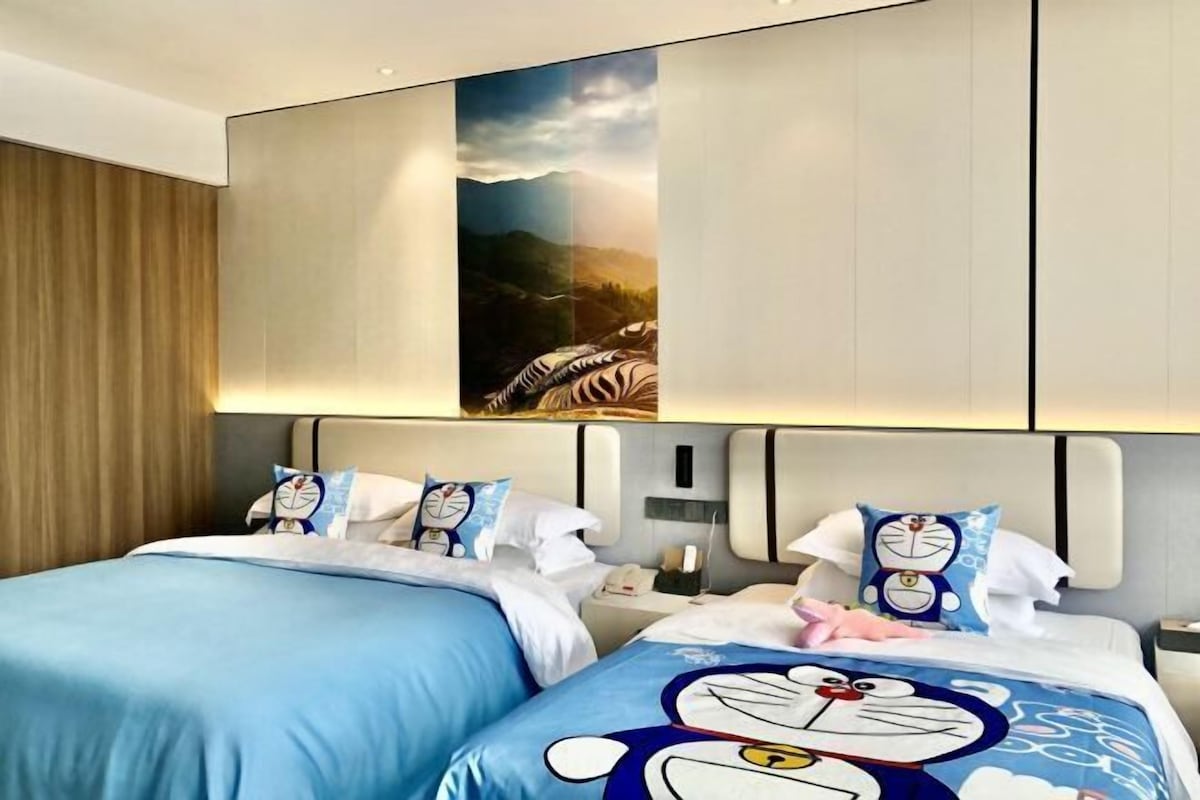 Habitación de hotel con dos camas, cada una con una colcha azul adornada con personajes de dibujos animados. Encima de las camas hay un cuadro grande que representa un paisaje montañoso.