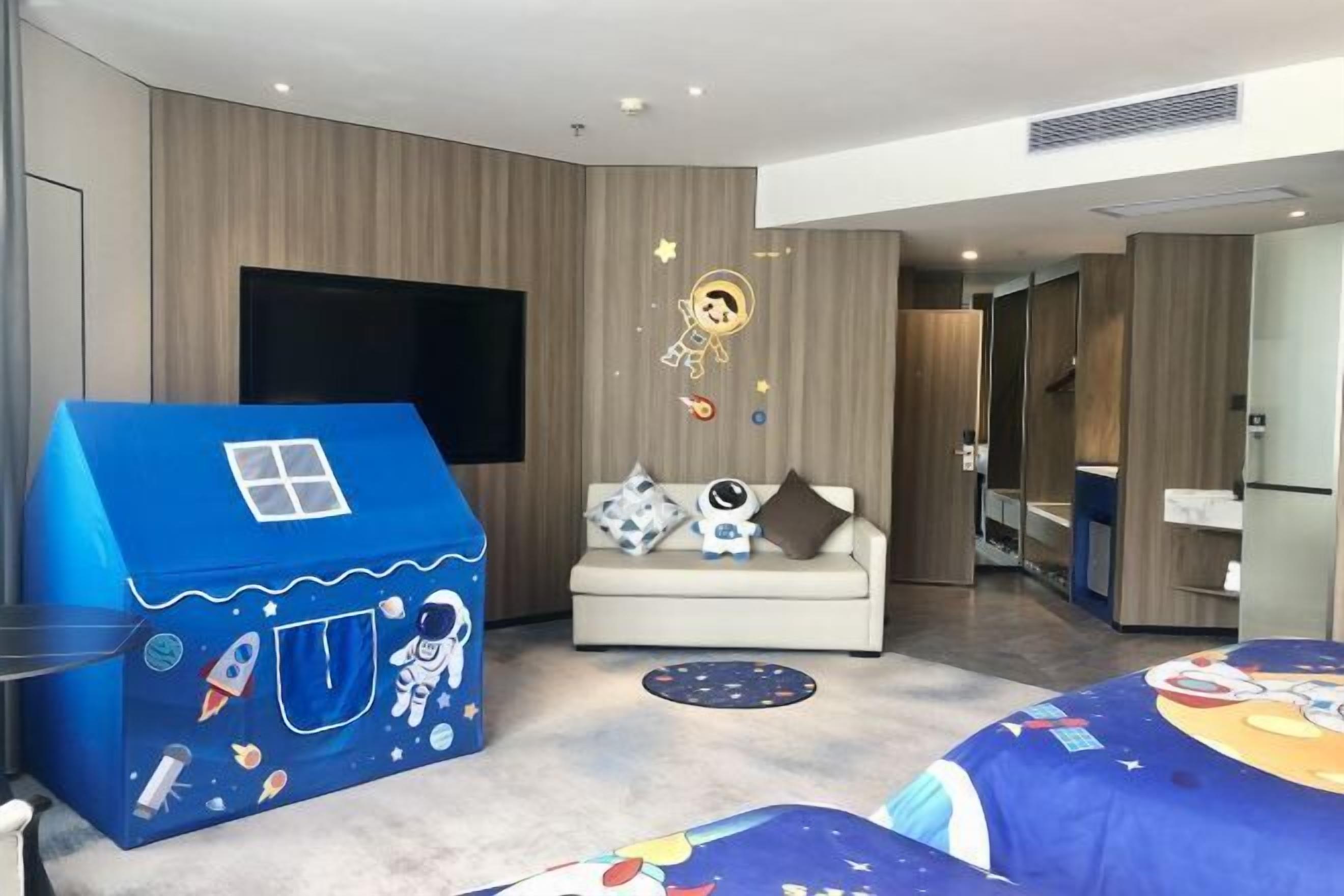 A modern hotel room with a large TV, a sofa, a blue playhouse, and a bed with a space-themed bedspread.
