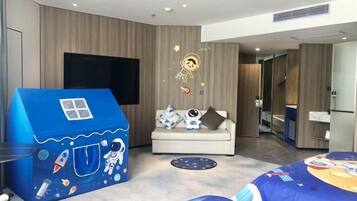 A modern hotel room with a large TV, a sofa, a blue playhouse, and a bed with a space-themed bedspread.