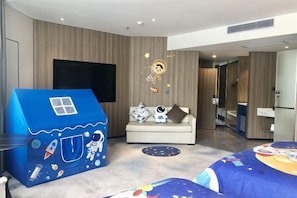 Una habitación de hotel moderna con un televisor grande, un sofá, una casita de juegos azul y una cama con una colcha de temática espacial.