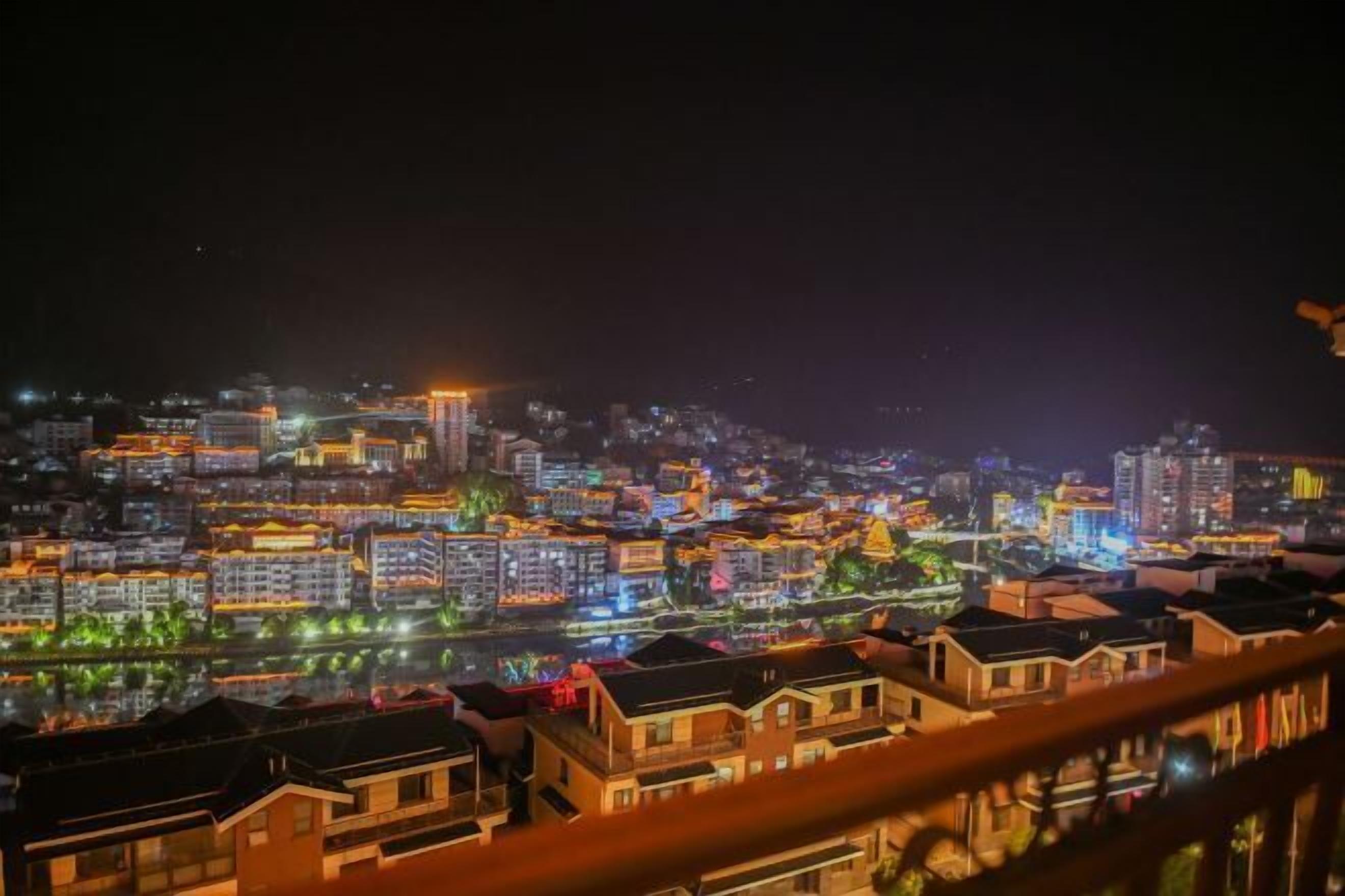 A nighttime cityscape with illuminated buildings and a river reflecting the lights.