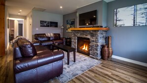 Una sala de estar con chimenea de piedra, sofás de cuero, un televisor de pantalla plana y un ventanal con vistas a un bosque.