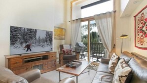 Een woonkamer met een televisie, leren bank, houten tv-meubel en uitzicht op een balkon met buitenmeubilair.
