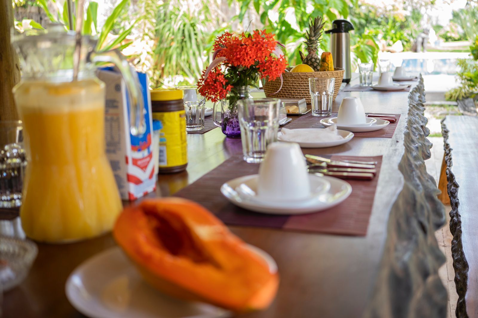 Une table de petit-déjeuner dressée avec des assiettes, un pichet de jus d’orange, un saladier de fruits et un panier de pain.