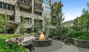 A stone building with multiple balconies, a circular outdoor seating area with a fire pit, and lush greenery. - The Lion Vail 2 Bedroom (Vail)