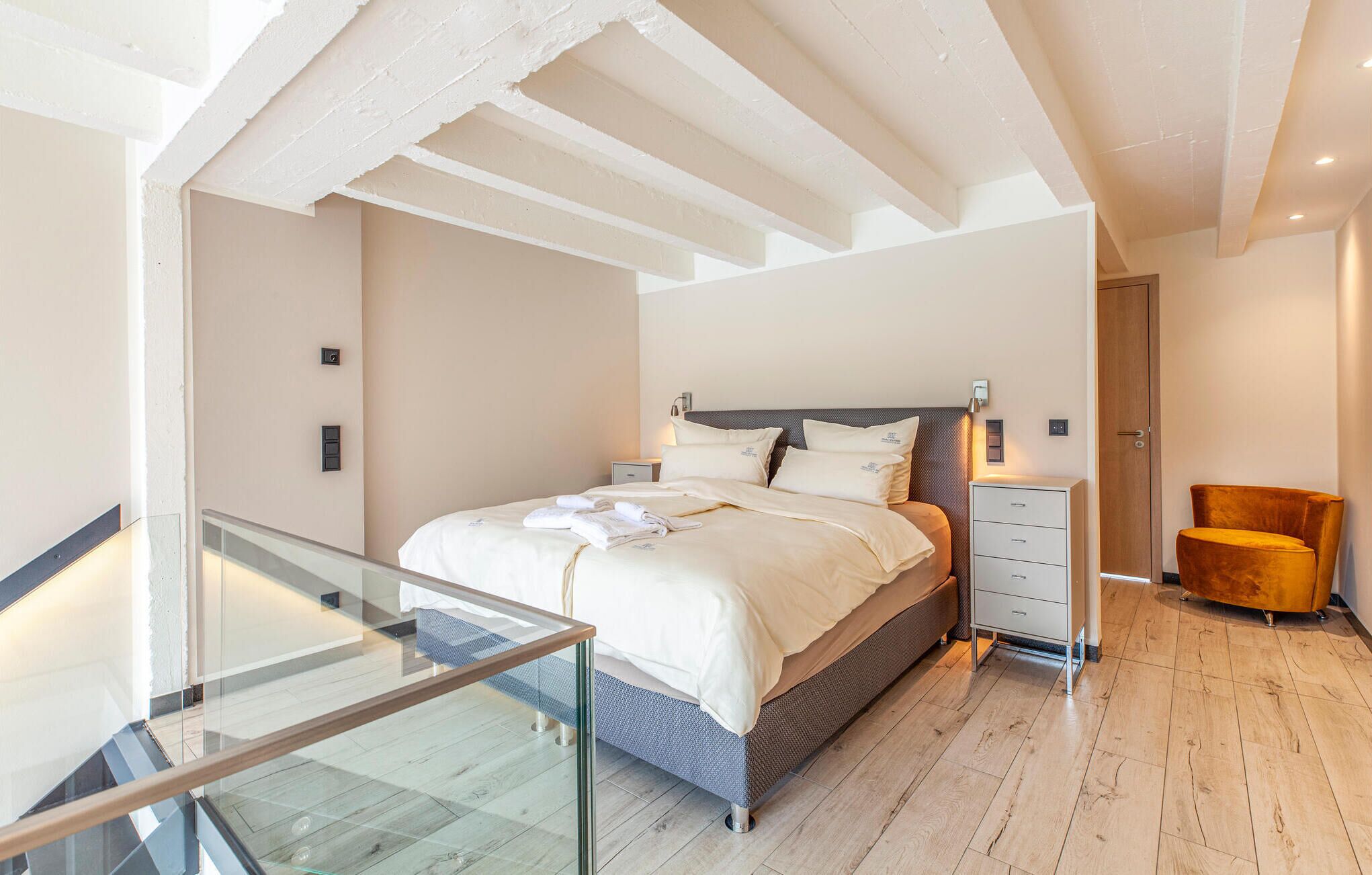 A modern bedroom with a glass balcony, a bed with white linens, a wooden chair, and a nightstand.