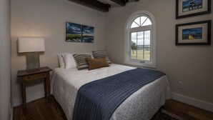A bedroom with a large bed, a nightstand with a lamp, a window with a view, and two framed pictures on the wall. - Comissary Middleburg (Huger)