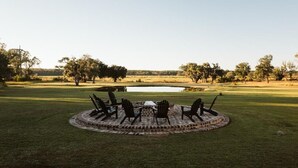 A circular outdoor seating area with chairs and a table, surrounded by a brick fire pit, set in a grassy field with trees and a pond in the background. - Comissary Middleburg (Huger)