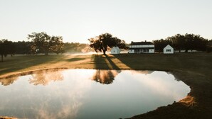 A serene landscape with a pond reflecting trees and buildings, surrounded by grassy areas and a clear sky. - Comissary Middleburg (Huger)