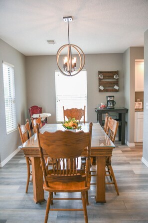 A dining room with a long table, six chairs, a chandelier, and a kitchen area with a coffee maker and a wooden board with handbags.
