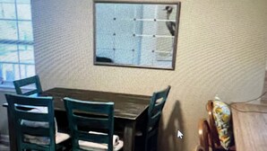 Un coin repas avec une table en bois et quatre chaises, un tableau d’affichage au mur et une fenêtre avec des stores.