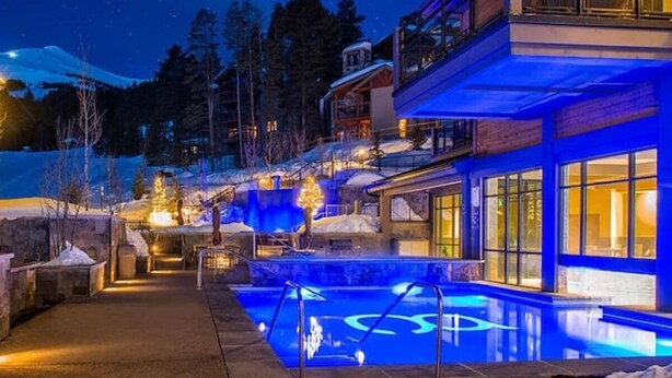 A nighttime view of a resort with a swimming pool, illuminated by blue lights, surrounded by snow-covered terrain and trees.