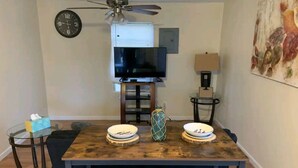 Un coin repas avec une table en bois, deux bancs, un téléviseur posé sur un meuble, une horloge et un tableau accroché au mur.