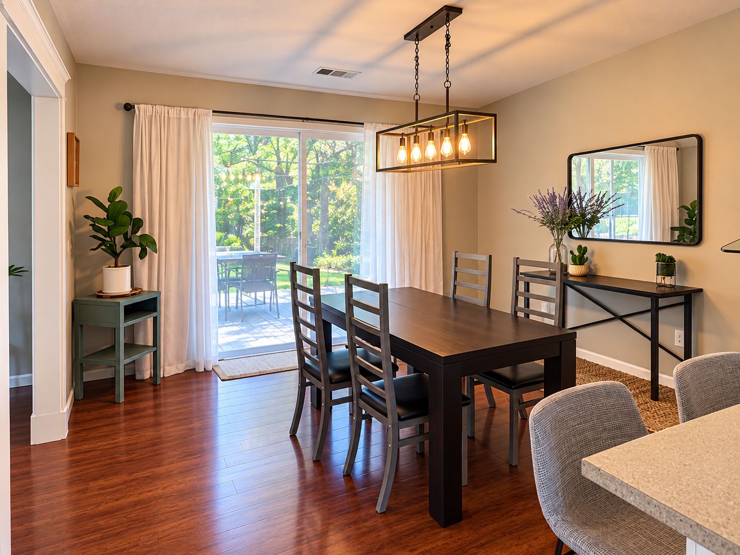 A dining area with a wooden table, chairs, a hanging light fixture, a potted plant, and a view through a glass door to an outdoor patio.