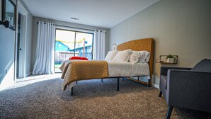 Ein Schlafzimmer mit Bett, Stuhl, einem Fenster mit Vorhängen und Blick auf ein Haus.