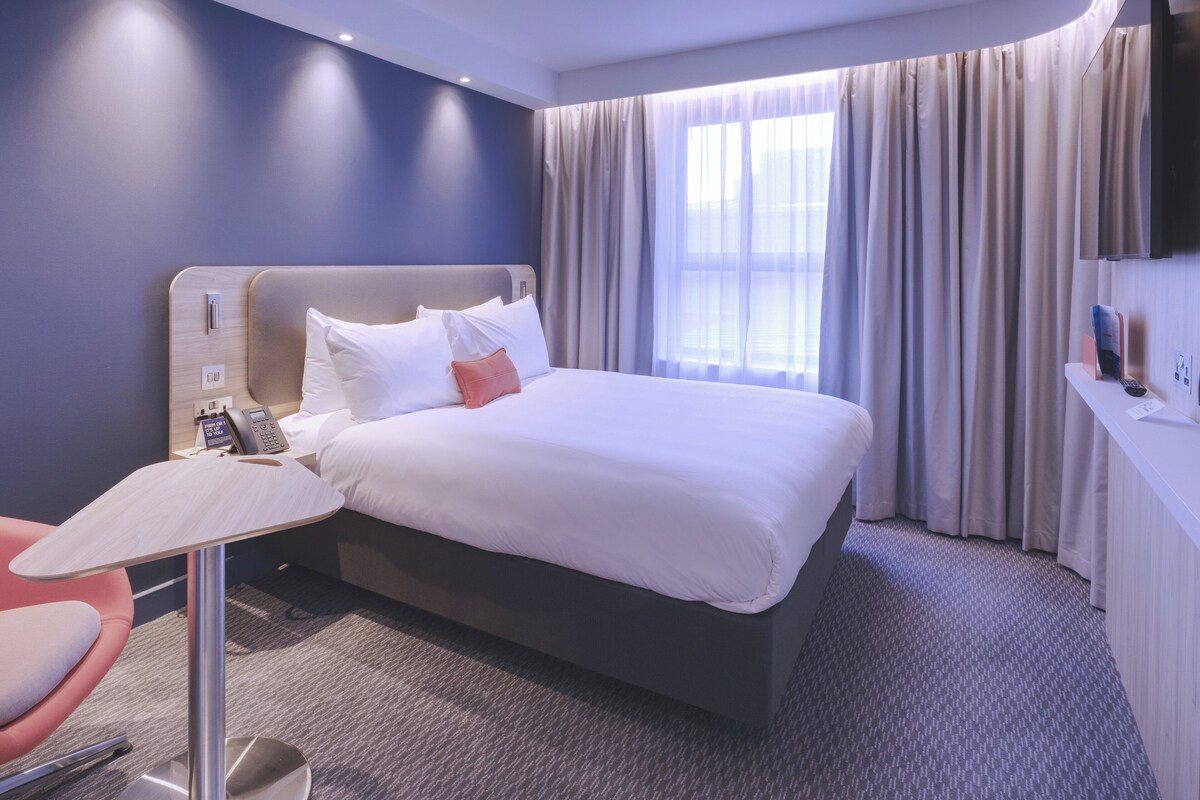 A hotel room with a bed, a desk, a chair, a TV, and a window with curtains.