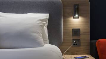 Una cama bien tendida con sábanas blancas, una mesita de noche con un teléfono inteligente cargando y una lámpara de pared.