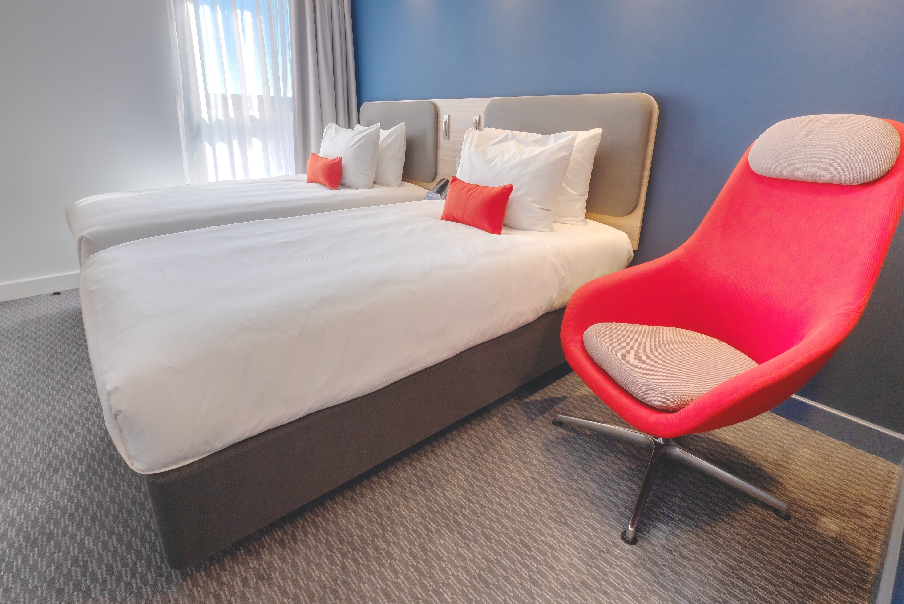 A hotel room with two beds, a red chair, and a view of the outdoors.