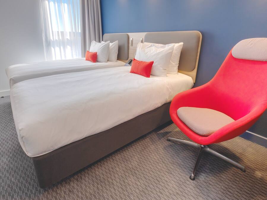 A hotel room with two beds, a red chair, and a view of the outdoors.
