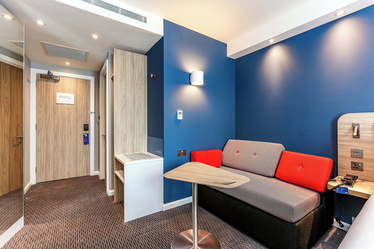 A modern hotel room with a blue accent wall, a grey sofa with red cushions, a small wooden table, and a built-in desk with a telephone.
