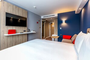 A modern hotel room with a large bed, a mounted TV, a small table with a chair, and a red accent wall.