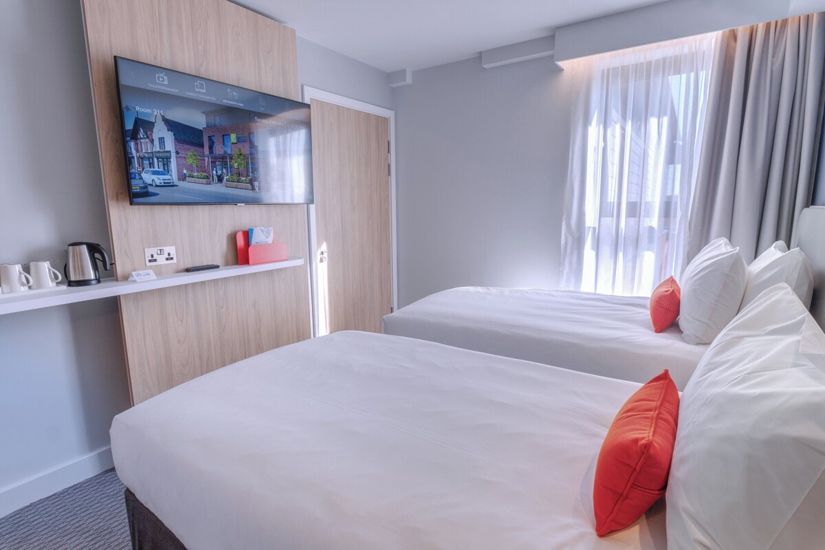 A hotel room with two beds, a TV mounted on the wall, a coffee machine, and a window with curtains.