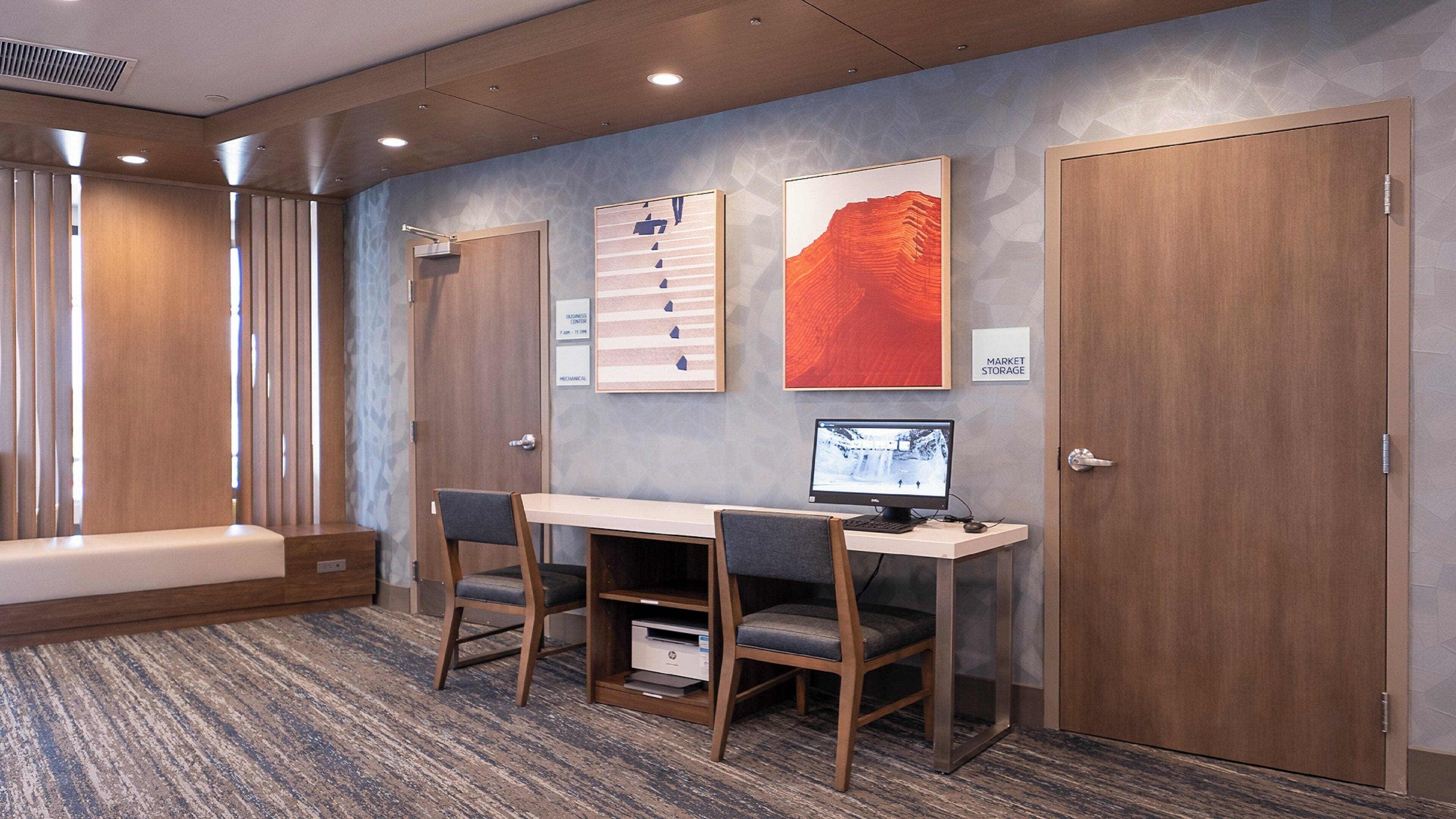 A hotel room with a desk, two chairs, a computer, and a large window with blinds.