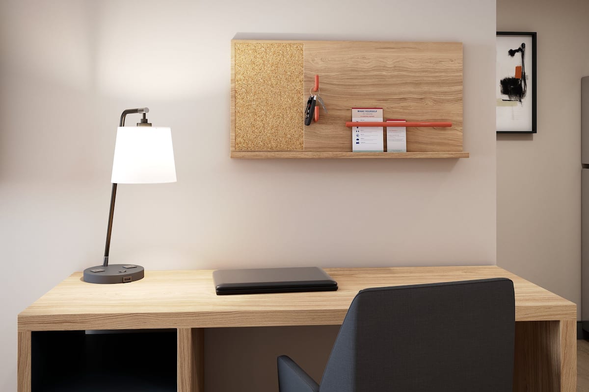 Ein moderner Schreibtisch mit einem Laptop, einer Schreibtischlampe, einem Korkbrett mit Schlüsseln und einem Namensschild sowie einem Bilderrahmen mit einem abstrakten Gemälde an der Wand.