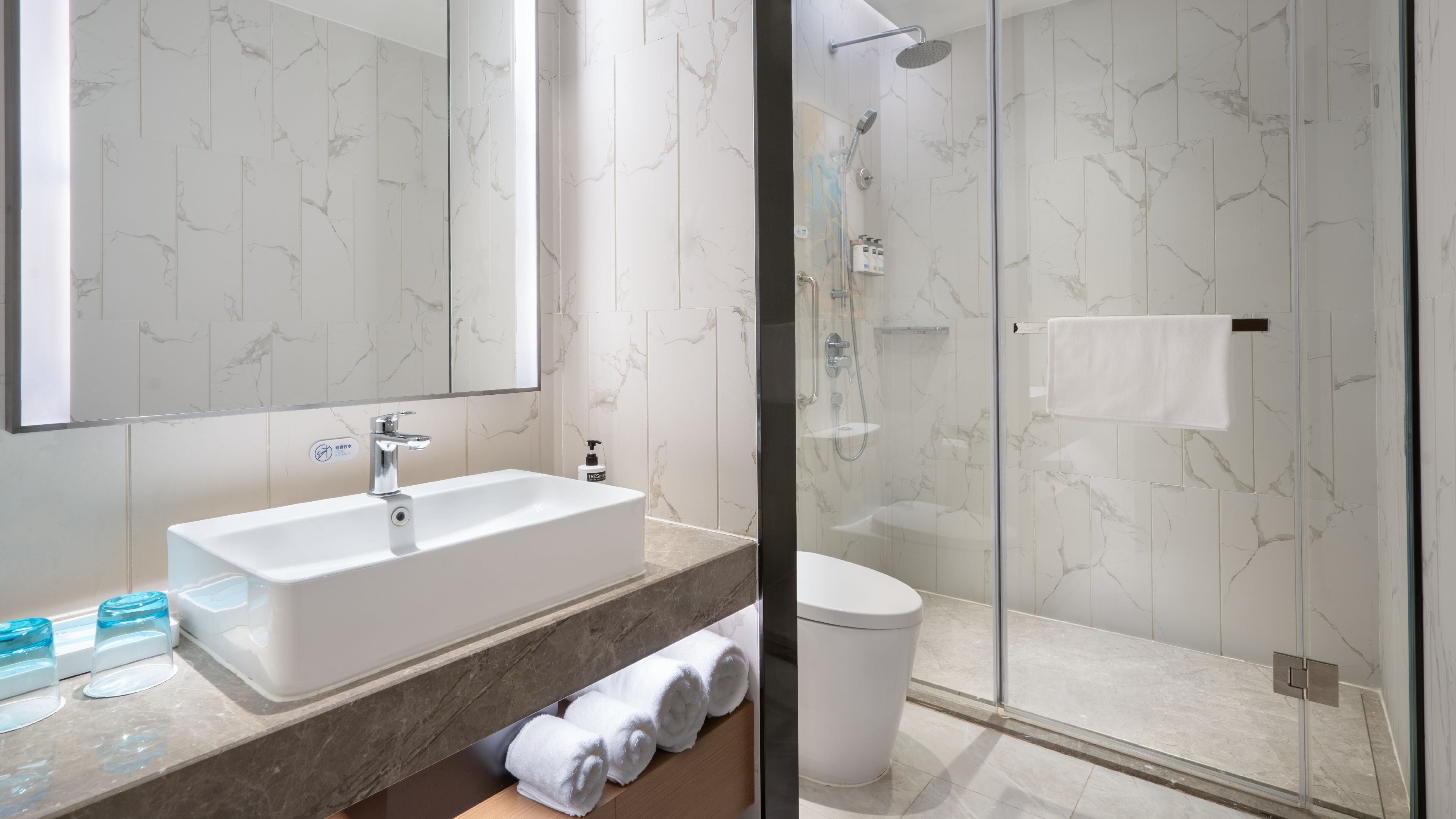Un baño moderno con un espejo grande, un lavamanos blanco, una ducha con mampara de vidrio y paredes con diseño de mármol.