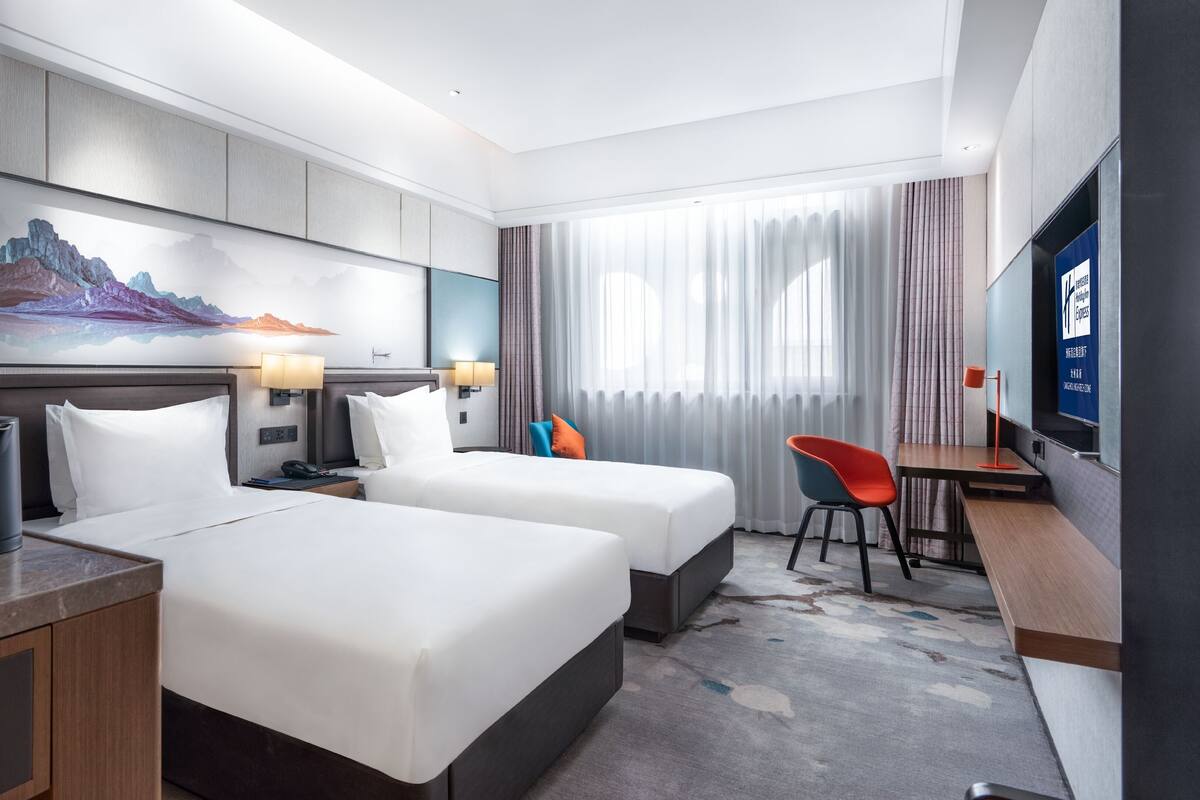 A hotel room with two beds, a desk, a chair, a TV, and a painting on the wall.