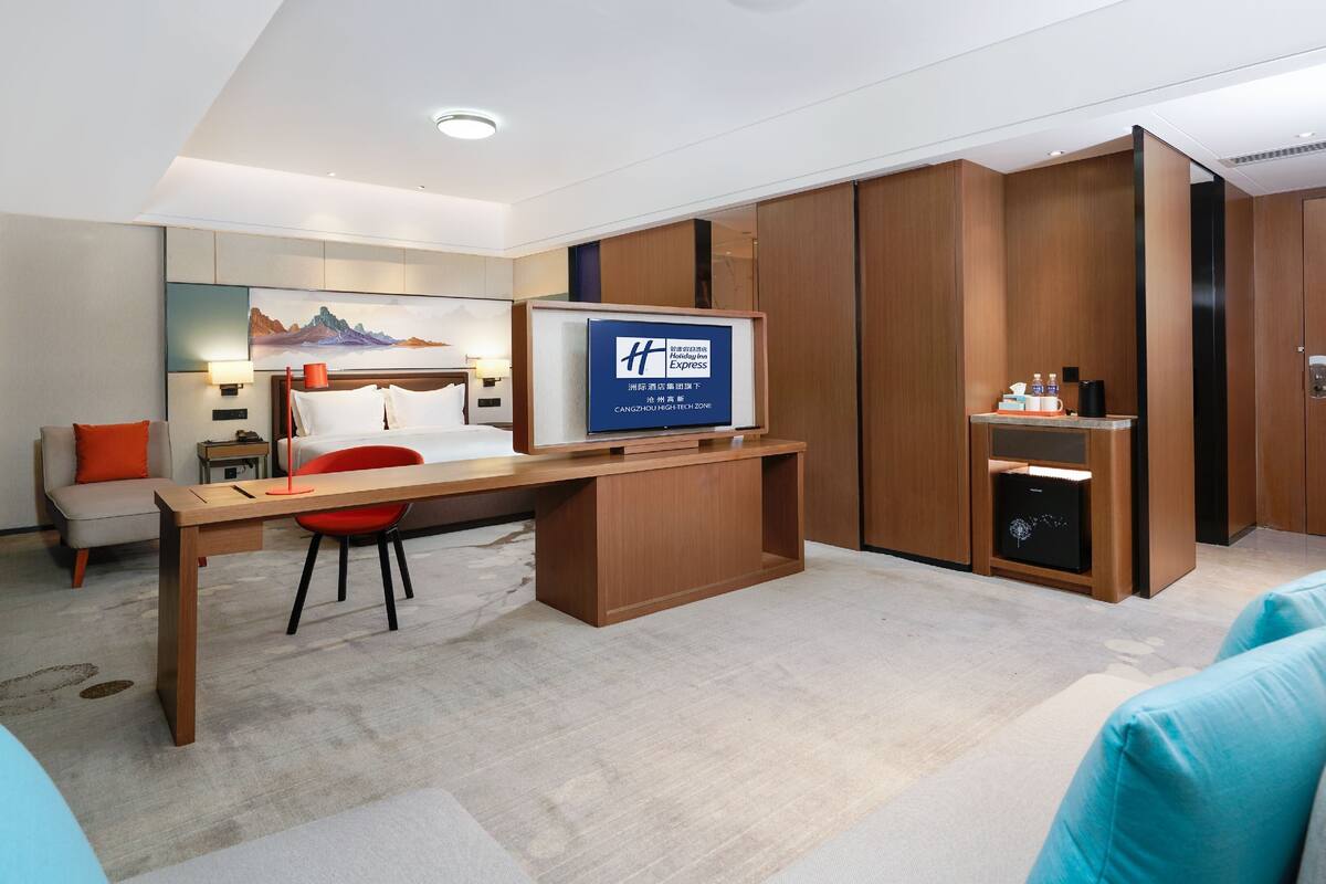 A modern hotel room with a wooden desk, a red chair, a bed, and a TV displaying a Holiday Inn Express sign.