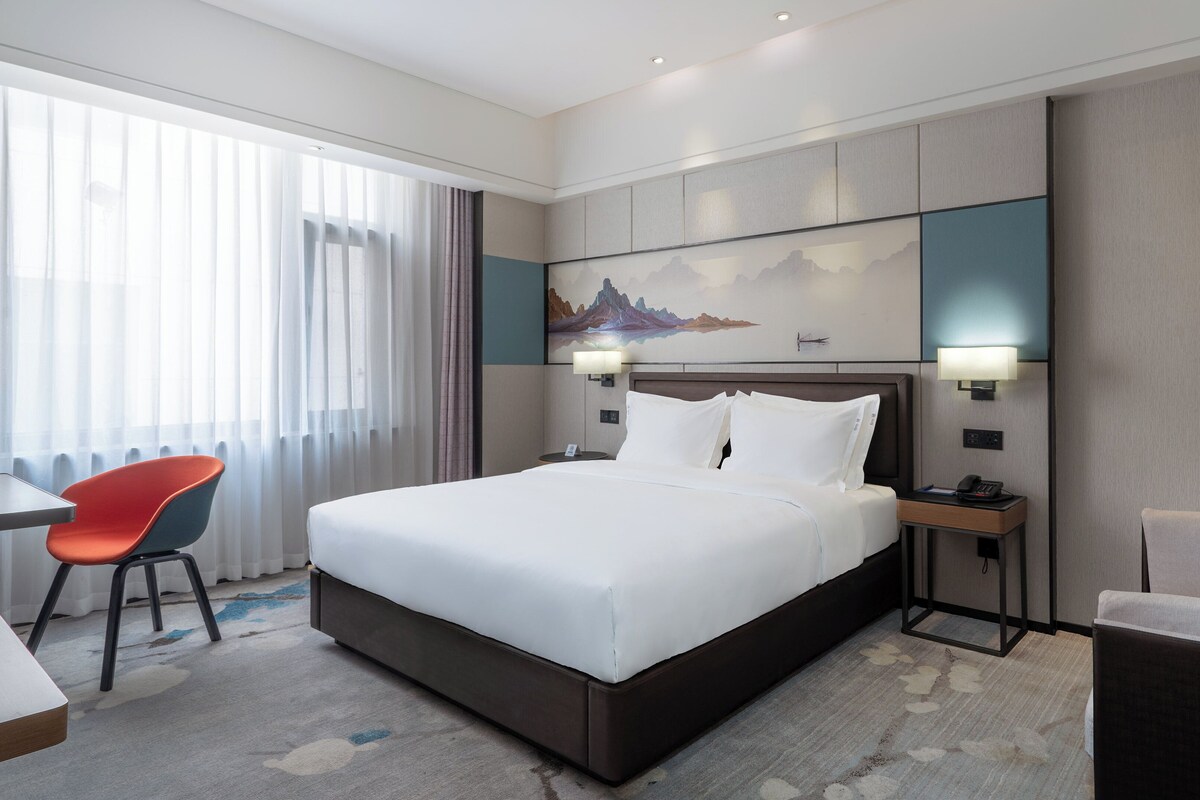 A hotel room with a large bed, a desk, a chair, and a painting on the wall.