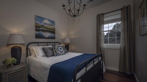 Een slaapkamer met een bed, twee nachtkastjes, een kroonluchter, een raam met gordijnen, en een schilderij aan de muur.