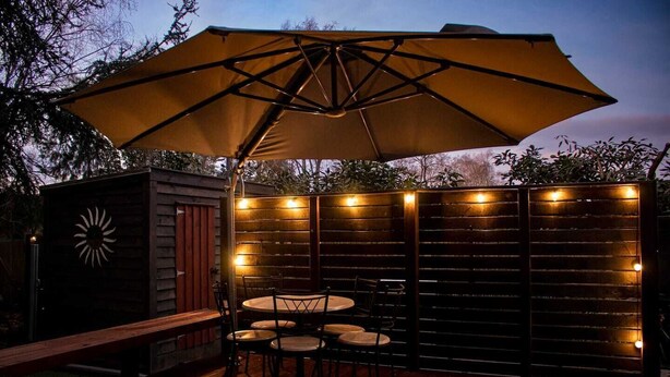 Een terras buiten met een houten tafel en stoelen onder een grote parasol, omringd door een houten omheining met lichtslingers.